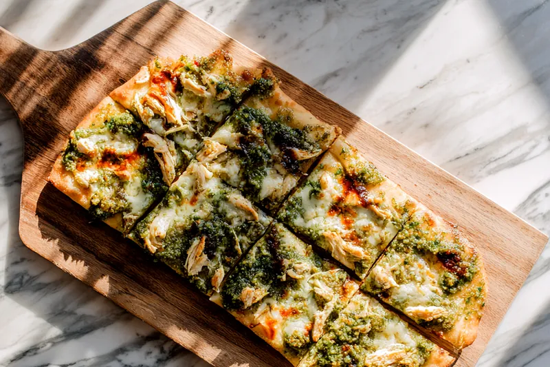 Side angle action shot of hands stretching pizza dough on floured wooden surface, with bowl of green pesto, shredded chicken, and cheese visible in background, natural kitchen lighting highlighting the texture of the dough