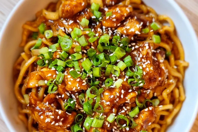 Fresh ingredients for sticky garlic chicken noodles including raw chicken pieces, garlic cloves, noodles, soy sauce, and honey arranged on a wooden cutting board